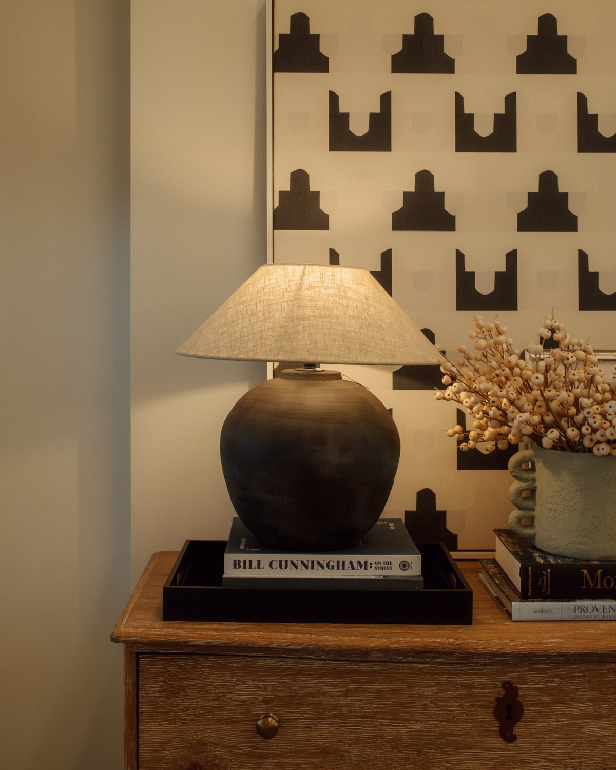 A black ceramic lamp with a linen shade on a weathered oak dresser in Waukee, set against black and cream patterned art.