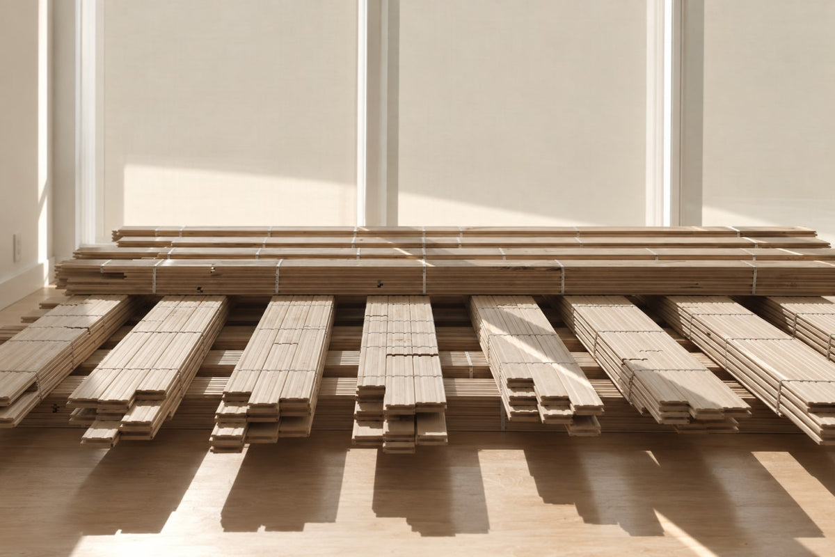 Bundles of unfinished hardwood flooring stacked on the subfloor of a Des Moines home under construction, acclimating in front of two tall covered windows.