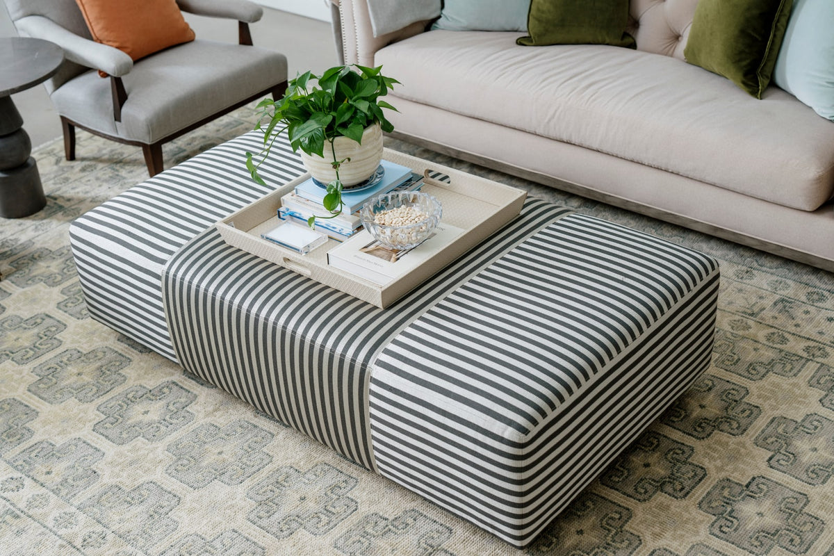 A living room in Waukee with a gray and white striped ottoman and a woven tray on a pale patterned wool rug.