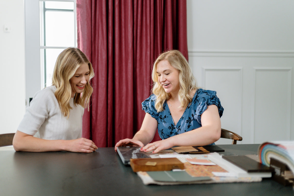 Two designers in a Waukee design studio laying out fabric and wood samples on a black table by deep red drapes.