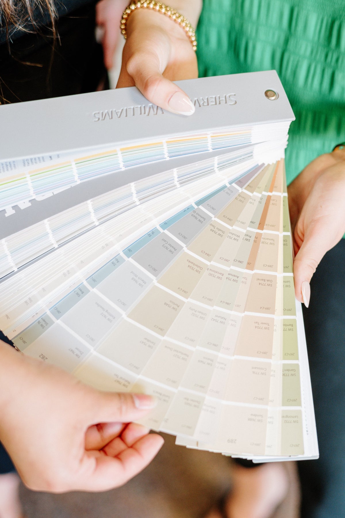 A designer holding open a Sherwin Williams paint fan deck on the warm neutral and soft green pages.