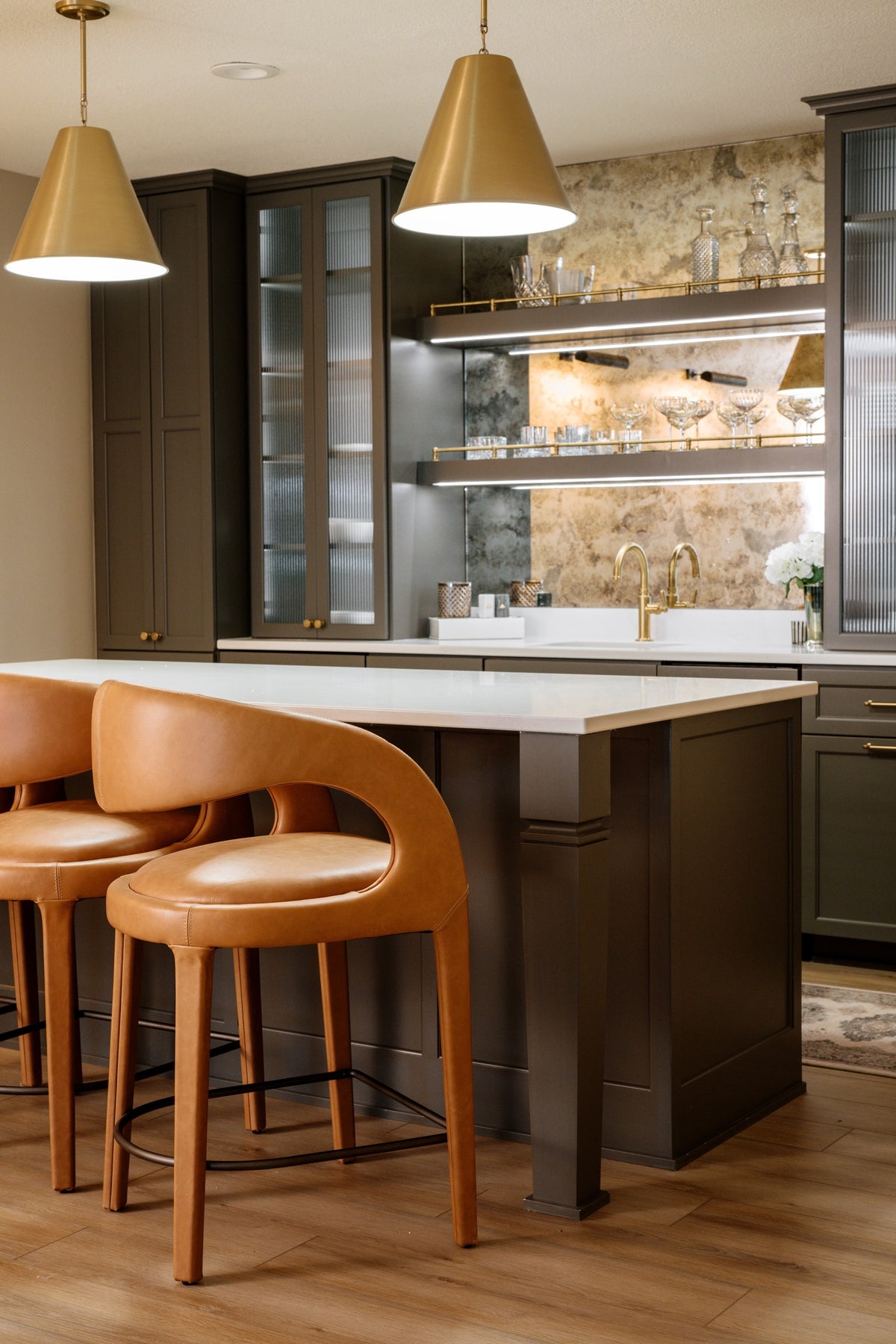 An olive green wet bar in Clive with an antiqued mirror backsplash behind lit brass rail shelves.